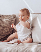 Ein Baby, das den leichten kurzen Babybody aus Musselinstoff trägt, sitzt auf einem Sofa.