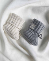 Zwei gestrickte Babyschuhe aus Cashmere mit Schnürung. Ein Schuh in creme, ein Schuh in hellgrau.