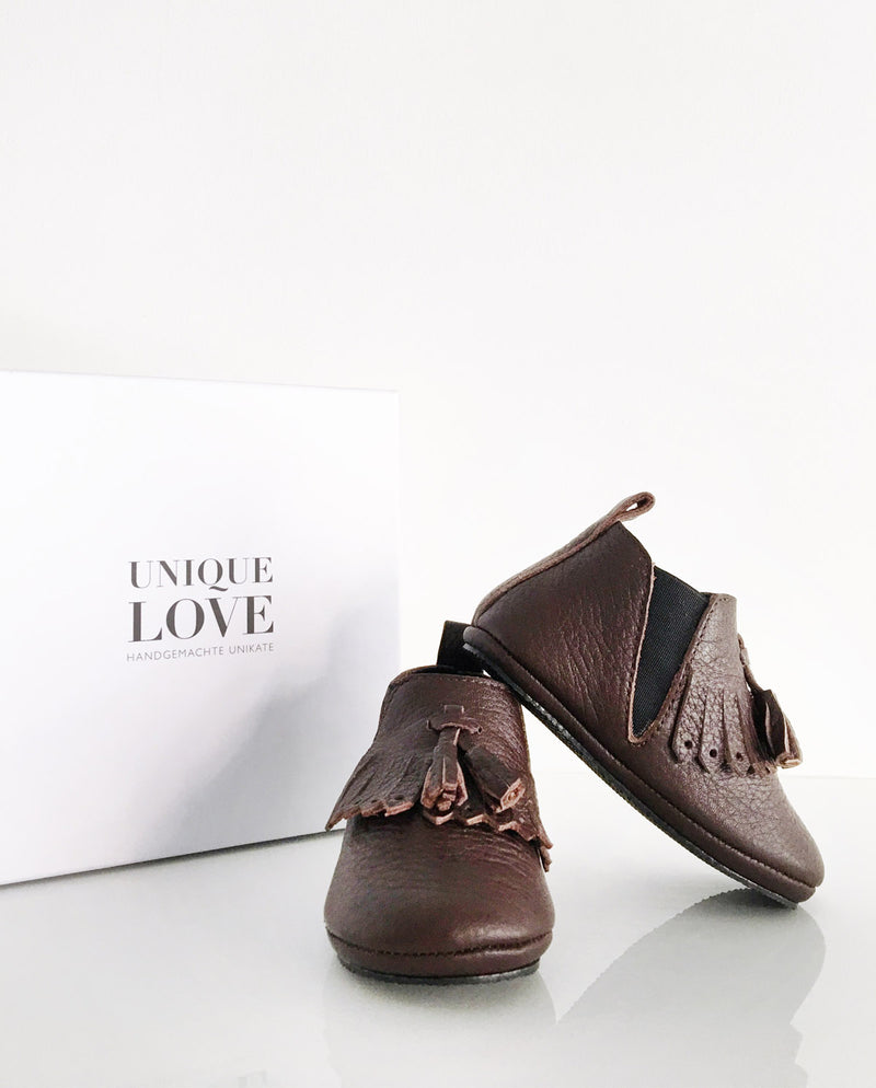 Chelsea Boots – Lederschuhe zum Laufen Lernen für Kleinkinder in der Farbe chocolate mit Fransen und zwei Quasten stehen vor einem Schuhkarton.
