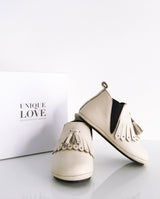 Chelsea Boots – Lederschuhe zum Laufen Lernen für Kleinkinder in der Farbe creme mit Fransen und zwei Quasten stehen vor einem Schuhkarton.