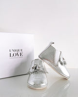 Chelsea Boots – Lederschuhe zum Laufen Lernen für Kleinkinder in silber glänzend mit Fransen und zwei Quasten stehen vor einem Schuhkarton.