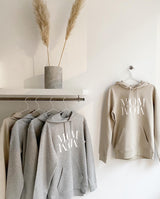 Drei graue und ein sand farbener Hoodie hängen auf Bügeln an einer Kleiderstange. Ein Kapuzenpullover in sand hängt an einem Haken an der Wand. Die Aufschrift MOM WOW ist klar zu erkennen.