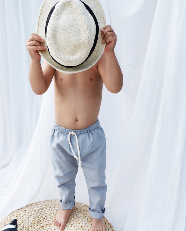 Kleiner Junge, der sich einen Hut vor das Gesicht hält, trägt die Leinenhose in hellgrau mit der Schnürung am Bund.