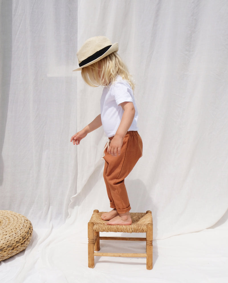 Ein Kleinkind mit Hut steht auf einem Holzhocker und trägt die Leinenhose in terrakotta.