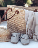 Ein Paar Moccasins Lederschuhe für Babys und Kleinkinder in der Farbe taube steht vor einer Strandtasche, in dem eine Kinder Badeponcho liegt.