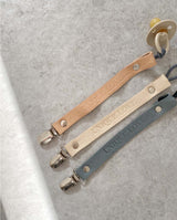 Drei Leder Schnullerbänder liegen auf einem Seidenpapier: Farben hellbraun, creme und grau.