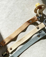 Sonnenstrahlen scheinen auf drei Leder Schnullerbänder in den Farben hellbraun, creme und grau. Am hellbraunen Band ist ein Schnuller in creme befestigt.