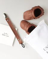 Setting aus Schnullerband aus Leder in der Farbe dunkelbraun mit geprägter Aufschrift „UNIQUE LOVE“, einer quadratischen Klappkarte "Hello Little one" und Moccasins in dunkelbraun.