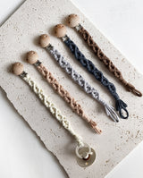 Kollektion aus fünf verschiedenen Farbtönen der Makramee Stoff Schnullerbänder: hellgrau, puder, creme, dunkelgrau, braun.