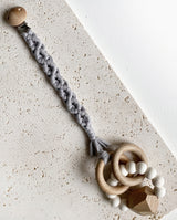 Schnullerband aus Stoff im Makramee Design mit Holzclip in der Farbe hellgrau mit einem Spielzeug Greifling daran.