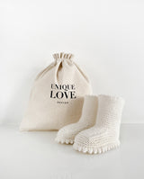 Ein Paar Strickschuhe für Babys mit Häkelkante in der Farbe creme steht vor einem Leinenbeutel mit Aufschrift "Unique Love Design".
