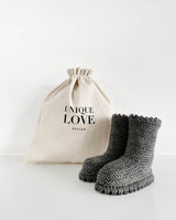 Ein Paar Strickschuhe für Babys mit Häkelkante in der Farbe dunkelgrau steht vor einem Leinenbeutel mit Aufschrift "Unique Love Design".