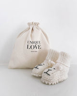 Ein Paar Strickschuhe für Babys in creme mit aufgestickten Löwengesichtern steht vor einem Leinenbeutel mit Aufschrift "Unique Love Design".