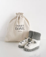 Ein Paar Strickschuhe für Babys in creme mit grauer Häkelkante und Bündchen und aufgestickten Löwengesichtern steht vor einem Leinenbeutel mit Aufschrift "Unique Love Design".