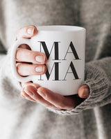 Zwei Hände umfassen die weiße Keramiktasse mit Henkel, auf der MAMA steht.