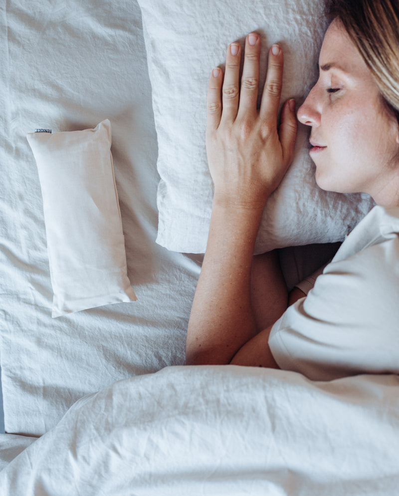 Frau schläft neben dem Augenkissen in creme, das auf Kopfhöhe neben ihr liegt, um so die Entspannung des Kissens zu spüren.