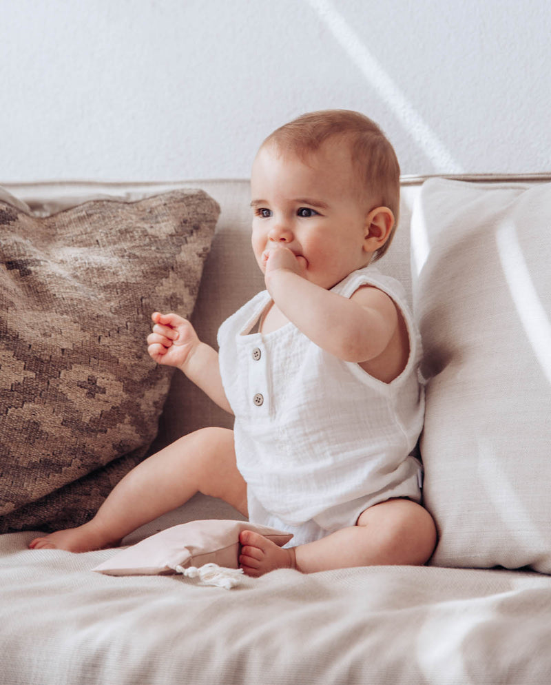 Ein Baby, das den leichten kurzen Babybody aus Musselinstoff trägt, sitzt auf einem Sofa.