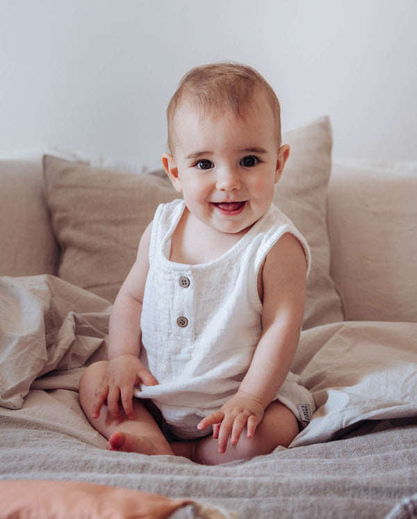 Ein Baby trägt den Musselinbody in creme und lächelt in die Kamera.