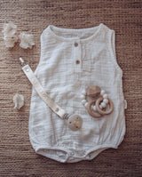 Musselinbody in creme für Babys liegt auf einem strukturierten Kordel-Untergrund zusammen mit einem Schnullerband in creme und einem Greifling/Beißring in weiß.