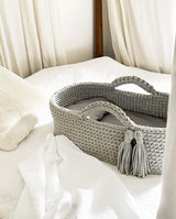 Babykörbchen aus gehäkelten Stofffäden in hellgrau steht auf einem Bett. Zwei Quasten als Detail sind am Henkel befestigt.