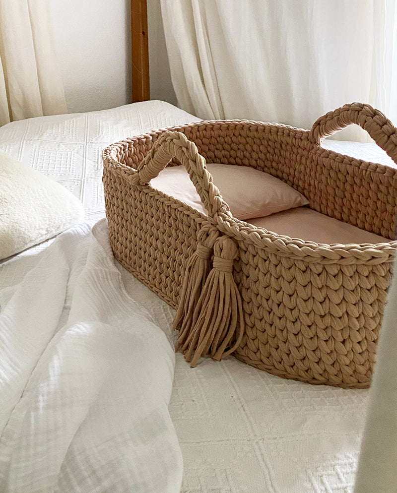 Babykörbchen aus gehäkelter Baumwolle in der Farbe Mocca steht auf einem Bett.