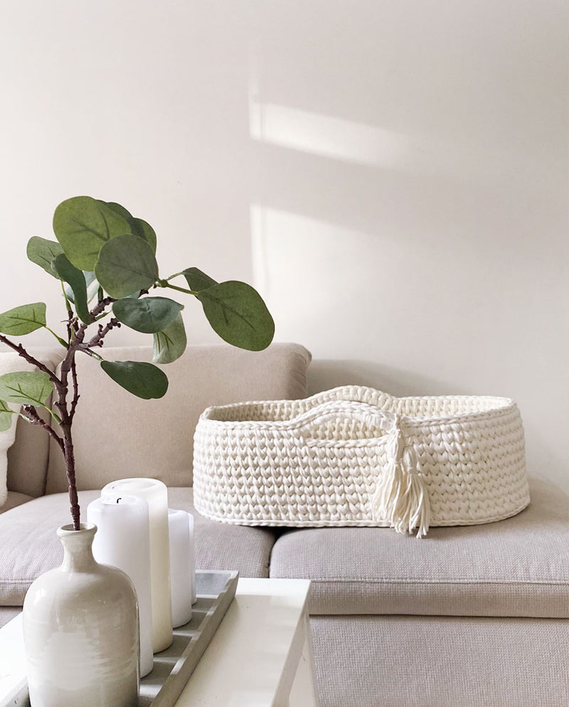 Gehäkeltes Babykörbchen in der Farbe cloud white mit zwei Quasten am Henkel als Detail steht auf einem Sofa.