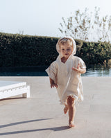 Junge läuft vom Pool in Richtung der Kamera. Er trägt den Sommer Badeponcho aus Baumwolle in der Farbe creme mit der Kapuze auf dem Kopf.