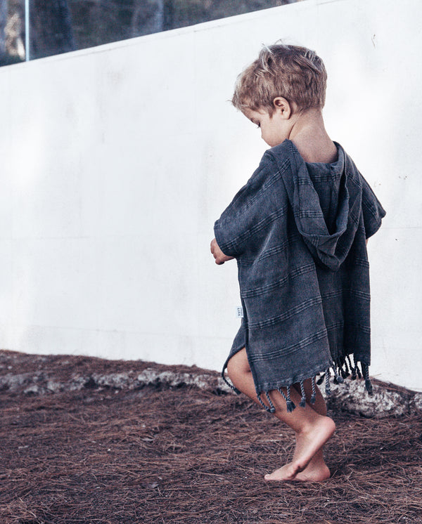 Junge trägt den Badeponcho in dunkelgrau mit Fransen am unteren Saum und Kapuze.