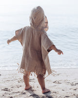 Ein Kleinkind steht am Strand mit ausgebreiteten Armen und trägt den Badeponcho im warmen beige-Ton mit Kapuze auf dem Kopf.