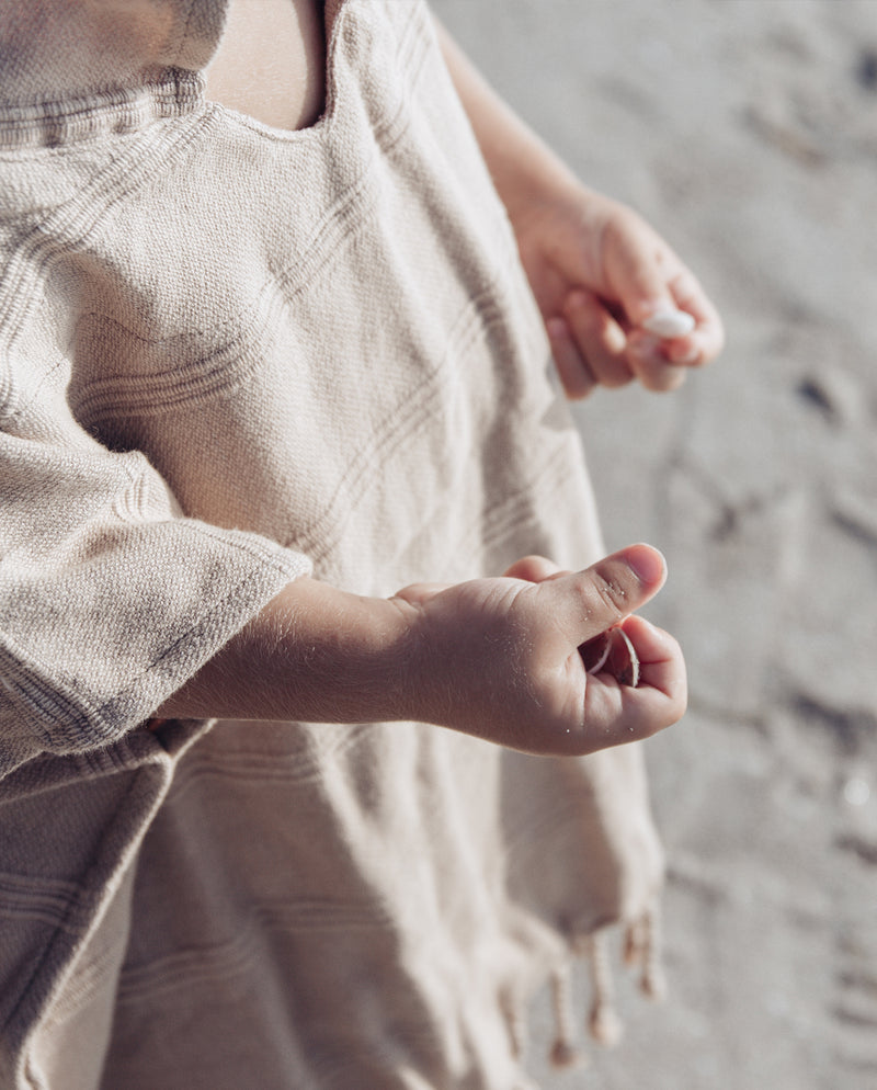 Kleinkind hält Muscheln und Sand in der Hand und trägt den Poncho für den Strand aus Baumwolle.