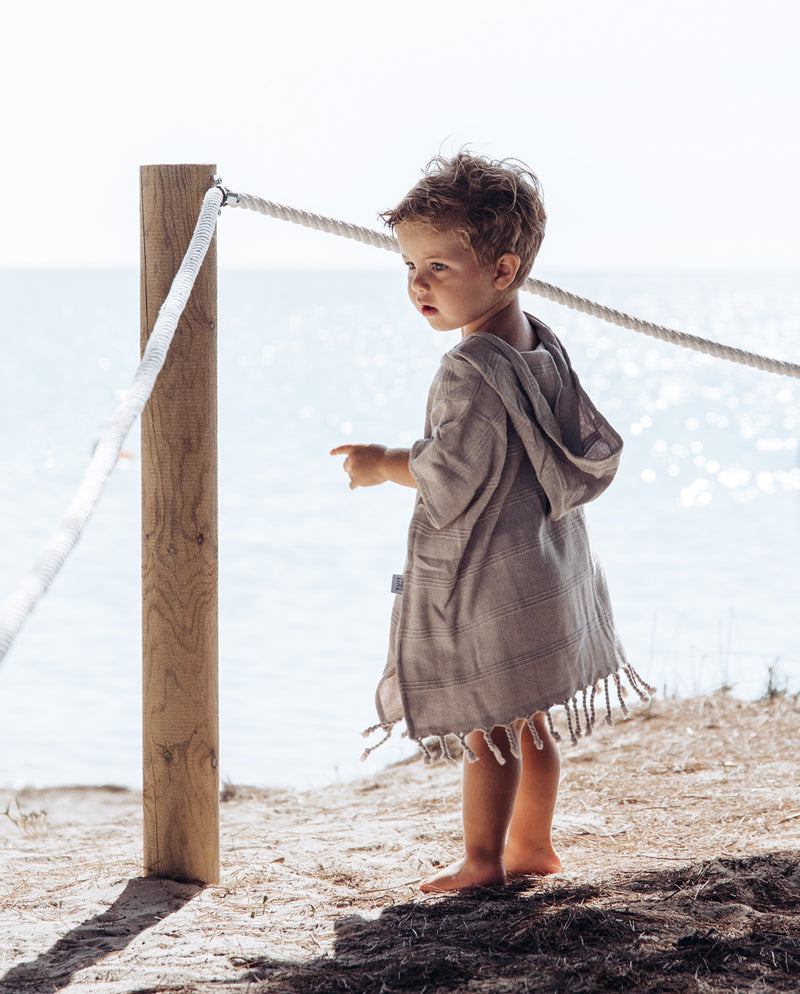 Ein kleiner Junge steht an einer Seilabsperrung am Meer, trägt den Badeponcho für Baby und Kleinkinder in sand und blickt zum Wasser.