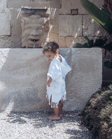 Kind steht an Brunnen in sommerlicher Umgebung und trägt den Badeponcho für Kinder in weiß mit Kapuze und Fransen am unteren Saum.