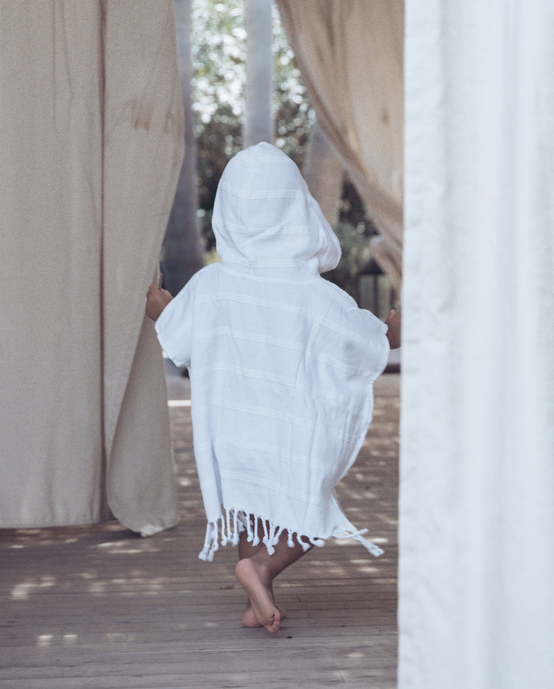 Kleinkind läuft bekleidet mit dem Hamam- Badeponcho für Kinder in weiß und der Kapuze auf dem Kopf auf einer sonnendurchfluteten Veranda.