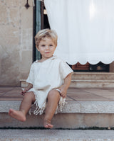 Ein fröhlicher blonder Junge sitzt auf der Terrasse und trägt den Badeponcho in creme.