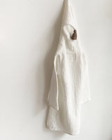 Musselin Bademantel in creme mit Quaste als Detail an der Kapuze hängt vor einer weißen Wand.