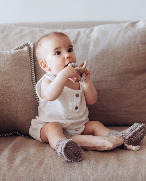 Baby sitzt auf einem Sofa und nimmt einen Beißring aus Holzringen und hellgrauen Silikonperlen in den Mund.