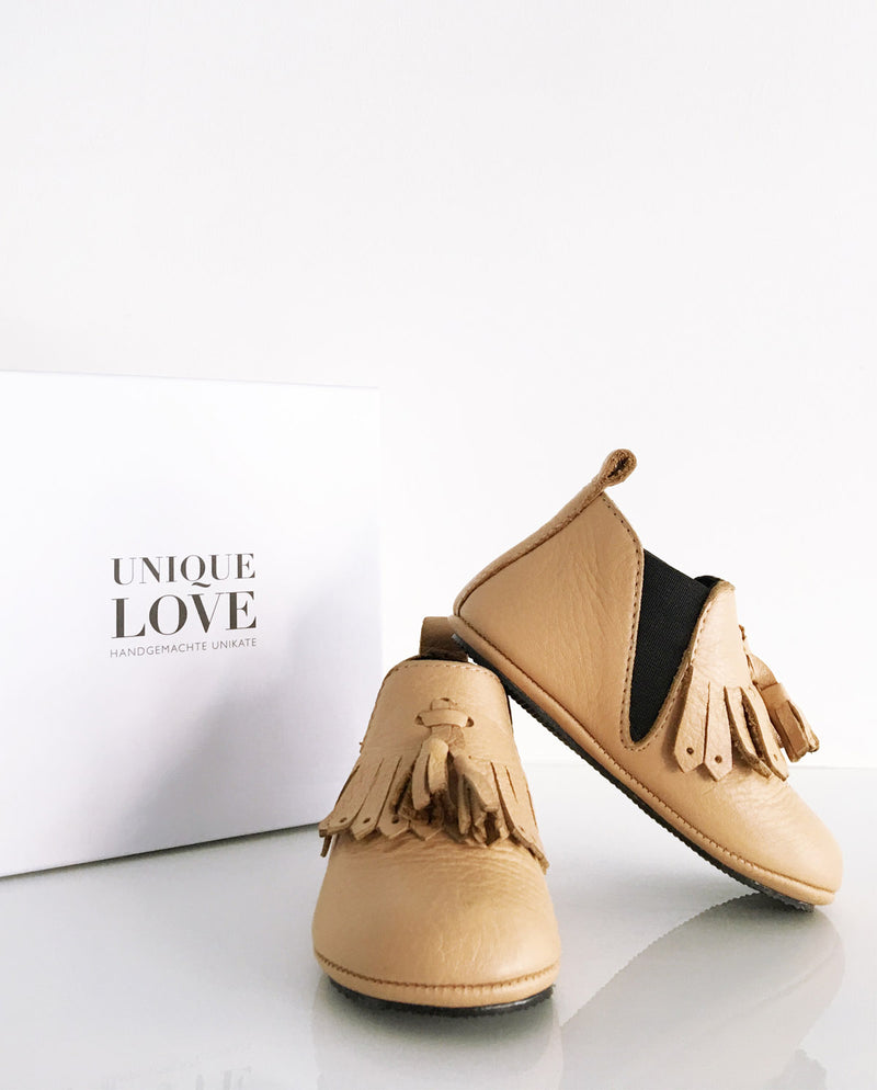 Chelsea Boots – Lederschuhe zum Laufen Lernen für Kleinkinder in der Farbe mocca mit Fransen und zwei Quasten stehen vor einem Schuhkarton.