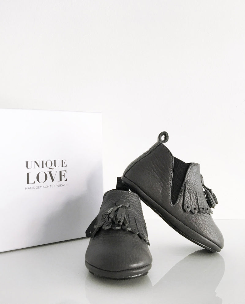 Chelsea Boots – Lederschuhe zum Laufen Lernen für Kleinkinder in der Farbe dunkelgrau mit Fransen und zwei Quasten stehen vor einem Schuhkarton.