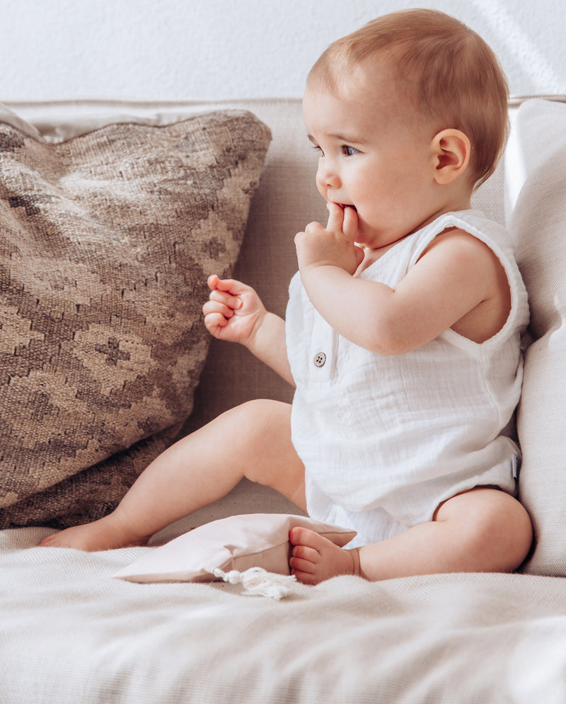 Baby sitzt auf einem Sofa und trägt den Musselinbody. Vor dem Kind liegt das Duftkissen mit Außenstoff in der Farbe Puder.