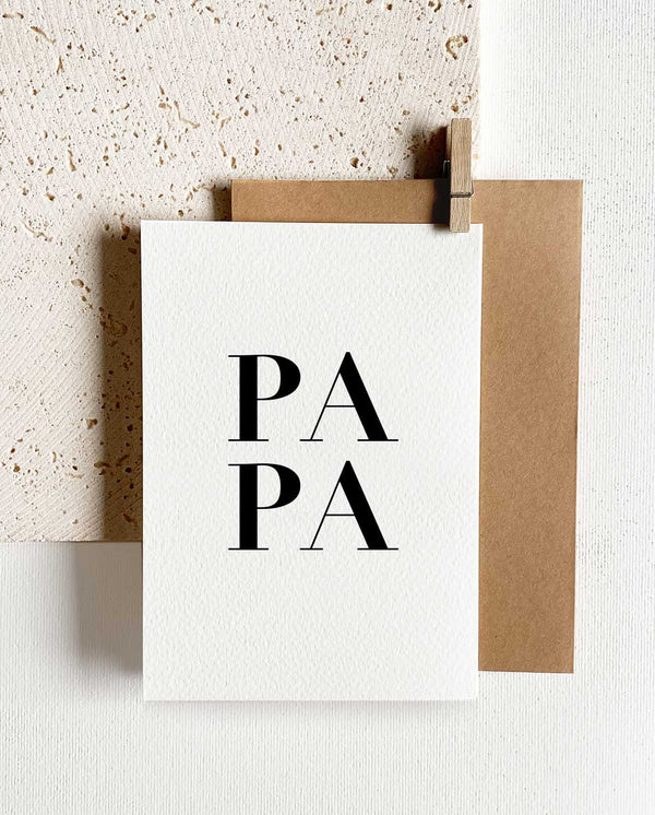 Rechteckige Grußkarte zum Klappen mit braunem Briefumschlag und Holzklemme. In schwarzer Schrift und in Großbuchstaben steht auf der Karte: "PAPA"