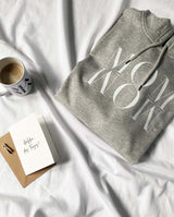 Ein gefalteter Hoodie MOM WOW in hellgrau liegt zusammen mit einer MAMA Tasse mit Kaffee und einer Karte "Heldin des Tages"