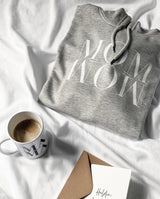 Nahaufnahme eines gefalteten Hoodies MOM WOW in hellgrau liegt zusammen mit einer MAMA Tasse mit Kaffee und einer Karte "Heldin des Tages"