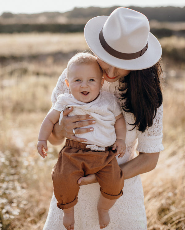 Eine Frau mit Hut hat ein Baby auf dem Arm, das in die Kamera lächelt und die Pumphose aus Leinen in der Farbe terrakotta trägt.