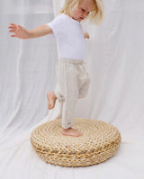 Ein Kleinkind steht auf einem Bein auf einem runden Holzkissen und trägt eine Leinenhose in creme.