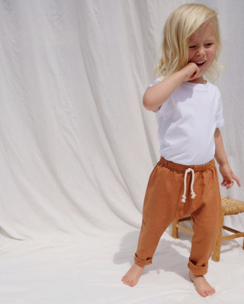 Ein Kleinkind steht vor einem weißen Tuch und präsentiert die Leinenhose terrakotta mit umgekrempelten Hosenbeinen.