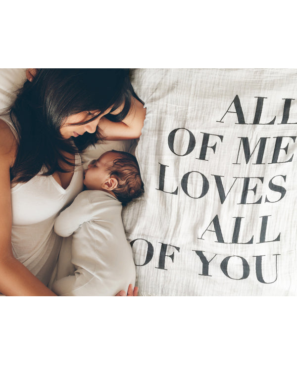 Eine Mama liegt mit ihrem Baby auf einem ausgebreiteten Musselintuch. Rechts neben ihnen ist der Schriftzug "All of me loves all of you" zu sehen.