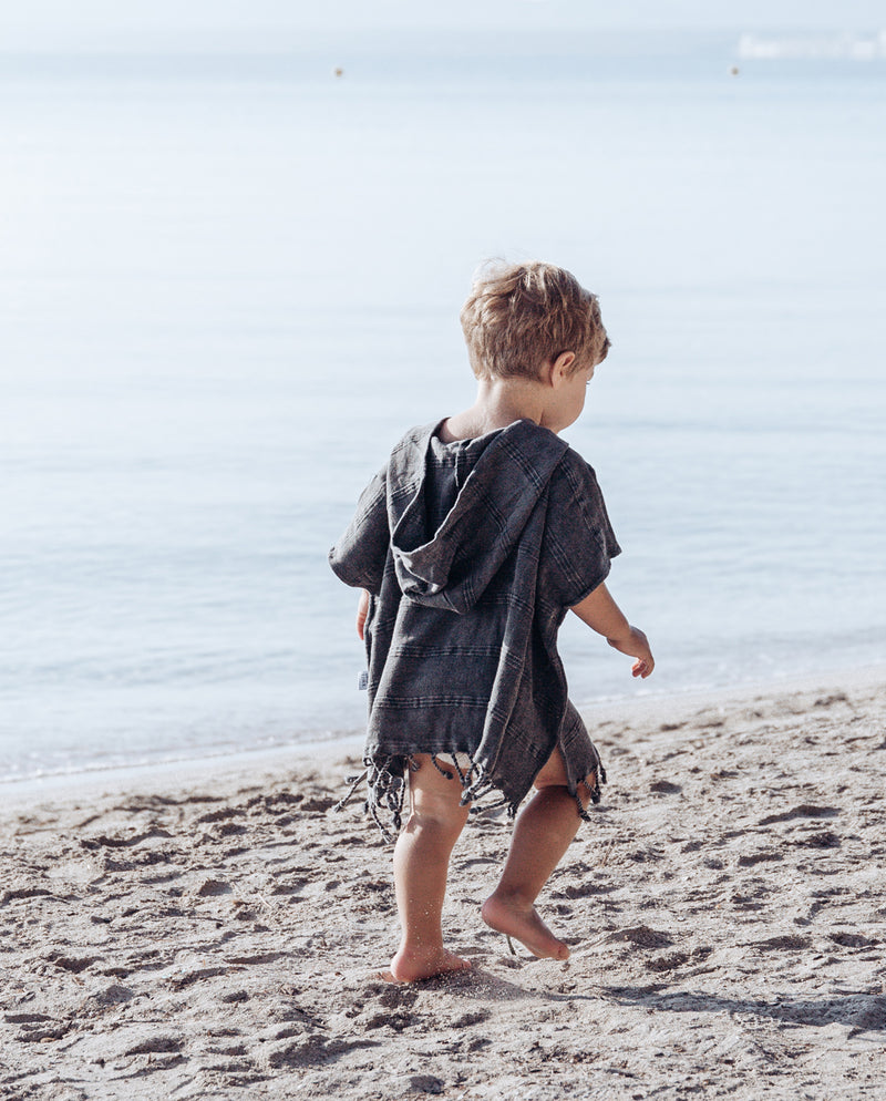 Kind läuft durch den Sand am Meer und trägt den Strandponcho in dunkelgrau mit Kapuze.