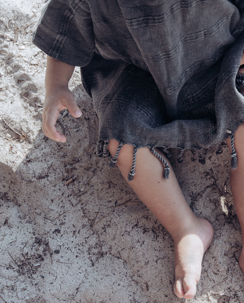 Kleinkind sitzt im Sand und trägt de dunkelgrauen Badeponcho mit Fransen am unteren Saum und einer mit uniquen Struktur des Hamam-Stoffes.