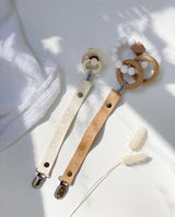 Moodbild zweier Schnullerbänder aus Leder in creme und hellbraun jeweils mit einem Schnuller und einem Baby-Spielzeug daran.