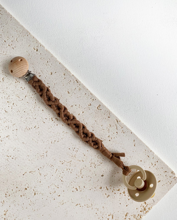 Schnullerband aus Stoff im Makramee Design mit Holzclip in der Farbe dunkelbraun mit einem Schnuller daran.